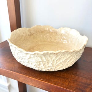 May include: A cream-colored ceramic bowl with a textured exterior that mimics the appearance of overlapping cabbage leaves. The bowl features a scalloped, wavy rim and a glossy finish, providing a distinct organic shape and a neutral tone.