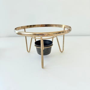 May include: A gold-toned metal stand featuring a circular top ring supported by three thin, angled legs. Suspended in the center of the structure is a small, dark-colored, cylindrical cup-shaped container made of a matte material.