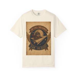 May include: An off-white short-sleeve t-shirt featuring a graphic of an owl wearing aviator goggles and a red scarf, flying a paper airplane. The design includes a full moon, wings, and the text ON-CALL SQUADRON and 3 AM NO SLEEP.