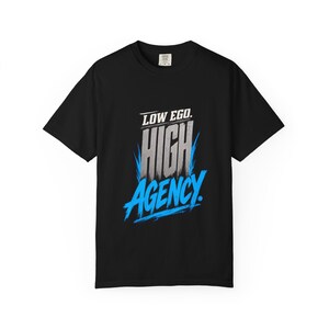 以下が含まれることがあります： 胸元にグラフィックプリントが施された黒の半袖Tシャツ。テキストは白で「LOW EGO」、メタリックシルバーのグラデーションで「HIGH」、鮮やかな青の筆致フォントで「AGENCY」と書かれており、文字の周囲にはダイナミックな青いアクセントが添えられています。