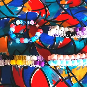 May include: Four handmade beaded bracelets featuring colorful plastic pony beads. One bracelet is circular with the text "TRANS" spelled out in letter beads. The other three are rectangular cuffs with horizontal stripes in white, black, purple, yellow, and pink.