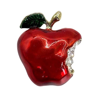 May include: A shiny red apple brooch with a gold-tone stem and a single green leaf. The apple features a bite mark on the right side, which is filled with small, clear, sparkling rhinestones that contrast against the smooth, glossy red enamel finish.