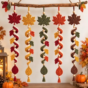 Op de afbeelding: Een wandhanger met vijf verticale macraméstrengen die aan een houten tak hangen. Elke streng bevat een groot gebreid esdoornblad, een spiraalvormig koord met kleine gebreide blaadjes in rood, geel, groen en bruin, houten kralen en een kwastje.