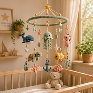 Könnte beinhalten: Ein hängendes Baby-Mobile mit gehäkelten Meerestieren, darunter ein blauer Wal, eine grüne Qualle, ein rosa Seepferdchen, eine grüne Schildkröte und ein gelber Fisch, ergänzt durch einen blauen Anker, Korallenformen und Sterne an einem grünen Ring.
