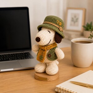 May include: A cream-coloured crocheted beagle figurine wearing a green bucket hat with a small yellow flower, a mustard yellow scarf with a green leaf detail, and a green vest with a brown button, standing on a round wooden base.