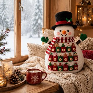 May include: A crocheted snowman advent calendar featuring a black top hat, a red and white striped scarf, and green mittens. The snowman has twenty small red and green pockets numbered one through twenty, each decorated with tiny festive ornaments.