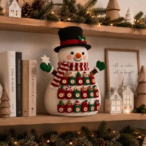 May include: A crocheted snowman advent calendar featuring a black top hat with a red band, a striped scarf, and green mittens. The snowman has twenty-four small numbered pockets in red and green, each decorated with tiny crocheted holiday ornaments.