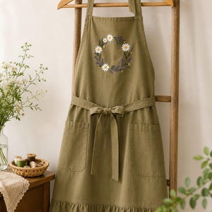 May include: An olive green apron featuring a central embroidered wreath of white daisies and purple lavender sprigs. The apron includes a tie-waist, two front pockets, a ruffled bottom hem, and adjustable neck straps tied in a bow at the top.
