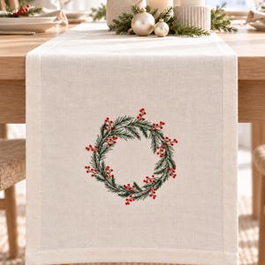 May include: A rectangular off-white table runner featuring a centered embroidered design of a circular wreath made of green pine needles and small red berries. The fabric has a visible woven texture with clean, straight stitched edges along the border.
