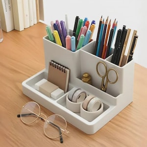 May include: A light grey plastic desk organiser with multiple compartments holding various pens, pencils, and markers. The organiser features a small spiral notebook, two rolls of washi tape, a pair of gold scissors, and a small eraser on the base.