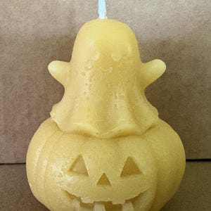 Peut inclure: Une bougie jaune en forme de fantôme assis sur une citrouille d'Halloween. Le fantôme a un petit visage souriant et les bras tendus, tandis que la citrouille présente des yeux triangulaires sculptés et une large bouche souriante.