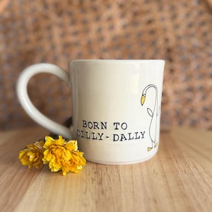 May include: A cream-coloured ceramic mug featuring a black line drawing of a goose and the text "BORN TO DILLY-DALLY" in black lettering. The mug has a curved handle and is decorated with a small illustration of a yellow-beaked goose.