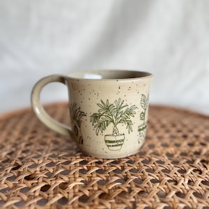 Puede incluir: Una taza de cerámica beige moteada con ilustraciones de líneas verdes de varias plantas en macetas. La taza tiene un asa curva en el lado izquierdo y un borde redondeado y liso. La superficie exterior está cubierta de pequeñas manchas marrones.