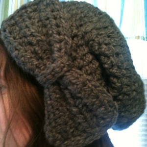 May include: A gray crocheted beanie with a large bow on top. The beanie is made with a thick yarn and has a slouchy fit.