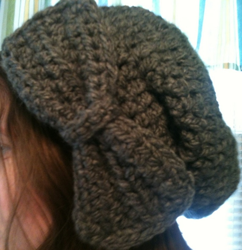 May include: A gray crocheted beanie with a large bow on top. The beanie is made with a thick yarn and has a slouchy fit.