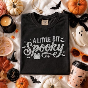Pode incluir: Uma t-shirt preta de manga curta com o texto branco A LITTLE BIT Spooky numa fonte decorativa. O design inclui pequenas ilustrações brancas de uma abóbora, estrelas e morcegos espalhadas em torno das letras centrais.
