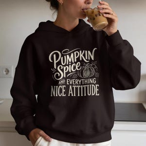 Könnte beinhalten: Ein dunkelbrauner Kapuzenpullover mit einem Grafikdruck in cremefarbenem Text, der Pumpkin Spice and Everything Nice Attitude lautet, begleitet von einer Strichzeichnung eines Kürbisses und dekorativen Schnörkeln.