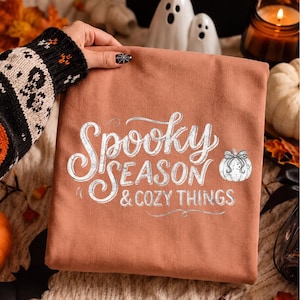 Pode incluir: Uma camisola de cor laranja queimado com o texto branco Spooky SEASON & COZY THINGS numa fonte decorativa e de bloco, acompanhada por um pequeno gráfico de uma abóbora branca com um laço, centrado na parte da frente.