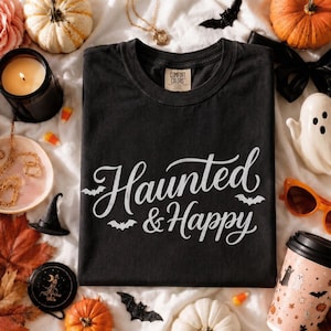 Pode incluir: Uma t-shirt preta de manga curta com gola redonda, apresentando o texto em caligrafia branca Haunted & Happy com pequenos gráficos de morcegos. A peça possui uma etiqueta da marca Comfort Colors no interior da gola.