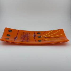 May include: A rectangular fused glass tray in a vibrant orange hue. The surface features a decorative pattern of small dark gray squares, thin yellow lines, and abstract purple accents, including a central cluster of intersecting lines.