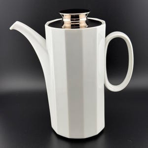May include: A white ceramic coffee pot with a faceted, hexagonal body, a long curved spout, and a large handle. The lid is topped with a polished, reflective metallic knob that contrasts with the matte white finish of the vessel's exterior.