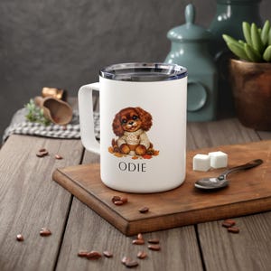 May include: A white cylindrical mug with a handle and a clear lid, featuring a graphic of a brown puppy wearing a beige sweater and holding a coffee cup, with the name ODIE printed in black capital letters below the illustration.