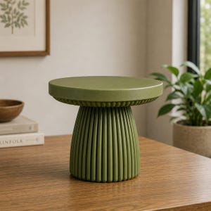 Puede incluir: Un soporte para pasteles de cerámica en color verde oliva mate que presenta una parte superior circular lisa y una base de pedestal ancha y estriada con crestas verticales. El soporte tiene un color sólido y un diseño estructurado y limpio.