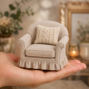 May include: A miniature beige armchair featuring a rounded back, rolled arms, and a ruffled skirt base. The chair includes a small, square, cream-colored throw pillow with a textured cable knit pattern, all crafted in a neutral fabric finish.