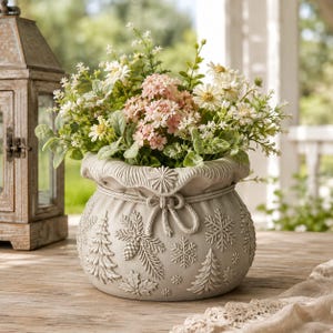 May include: A light grey ceramic planter shaped like a drawstring pouch, featuring embossed winter motifs including pine trees, snowflakes, and pinecones. The planter is filled with a bouquet of white daisies and small pink flowers.