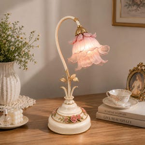 May include: A table lamp with a curved cream-colored stem and a base decorated with sculpted pink roses. The light fixture features a translucent, ruffled, pale pink glass shade shaped like a flower, supported by a gold-toned metal cap.