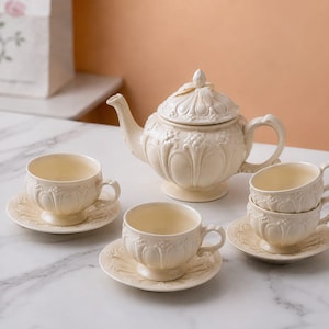 May include: A cream-colored ceramic tea set featuring a teapot with a lid and three matching teacups on saucers. Each piece is decorated with embossed floral and leaf patterns, a scalloped base, and a curved handle, all in a uniform matte finish.
