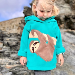 May include: A turquoise fleece hoodie featuring a brown sloth applique. The sloth is hanging upside down. The hoodie has a hood and long sleeves. The background is a rocky landscape.