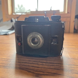 Op de afbeelding: Een vintage zwarte Ansco Clipper boxcamera met een getextureerde behuizing, een centrale ronde lensbehuizing met metalen rand, een opwindknop aan de bovenkant, een kleine zoeker en een rode cirkelvormige indicator op het voorpaneel.