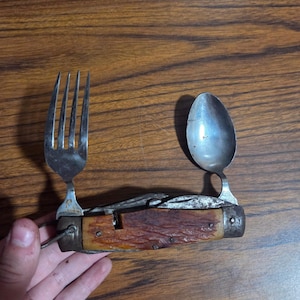 May include: A multi-tool camping utensil featuring a silver-toned metal fork and spoon extending from a textured, brown, bone-like handle. The folding tool includes a metal loop at one end and a weathered, metallic central body section.