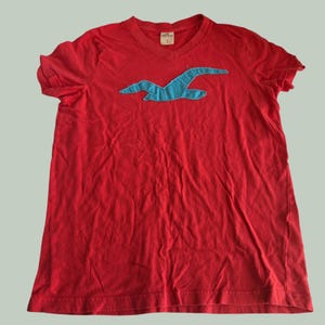 Könnte beinhalten: Ein leuchtend rotes Kurzarm-T-Shirt mit V-Ausschnitt. In der Mitte der Brust befindet sich eine große, aufgenähte hellblaue Möwengrafik. Ein kleines weißes Etikett im Kragen zeigt den Markennamen Hollister und die Größenangabe.