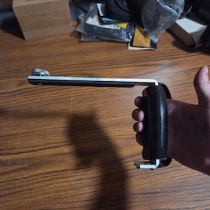May include: A black and silver camera flash bracket featuring an L-shaped metal frame with a black plastic grip handle and a silver mounting screw at the end of the horizontal arm for attaching a camera or lighting accessory.