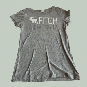 May include: A heather grey short-sleeve t-shirt featuring a white graphic of a moose silhouette next to the text "FITCH" in a bold, block font. Below the word "FITCH," the text "NEW YORK" is printed in a subtle, tonal grey font.