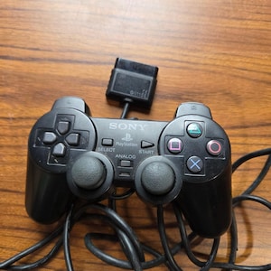 Puede incluir: Un mando de juego con cable Sony PlayStation de color negro que incluye dos joysticks analógicos, una cruceta, cuatro botones de acción con símbolos geométricos y botones centrales etiquetados como Select, Start y Analog, con cable.