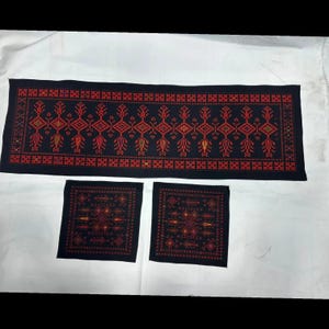 May include: A long rectangular black table runner and two matching square placemats, each featuring intricate cross-stitch embroidery in vibrant red, orange, and yellow geometric patterns arranged in repeating diamond and floral motifs.