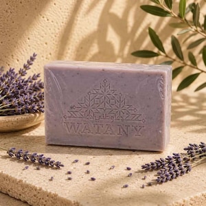 Puede incluir: Una pastilla de jabón rectangular de color lavanda claro con una superficie texturizada. El jabón presenta un patrón geométrico en relieve y el texto WATANY en letras mayúsculas. Pequeñas ramas de lavanda están esparcidas alrededor.