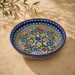 May include: A round ceramic bowl featuring a vibrant hand-painted floral pattern with red, blue, yellow, and orange flowers set against green foliage. The rim is decorated with a dark blue border containing white semi-circles and geometric lines.
