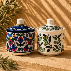May include: Two ceramic lidded jars with distinct hand-painted designs. The left jar features a vibrant floral pattern in red, blue, and yellow. The right jar displays a green olive branch motif on a white background, both with matching lids.