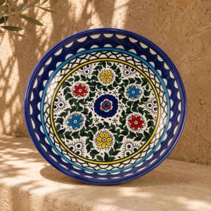 May include: A round ceramic bowl featuring a vibrant hand-painted pattern with a deep blue rim. The interior displays a central dark blue floral motif surrounded by alternating red, yellow, and blue flowers nestled within green leafy vines.
