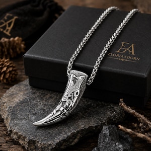 May include: A silver-toned pendant shaped like a curved tooth or tusk, featuring intricate engraved patterns and a polished tip. The pendant hangs from a silver-toned wheat chain. The piece is displayed near a black box with the text ELORIAADORN.