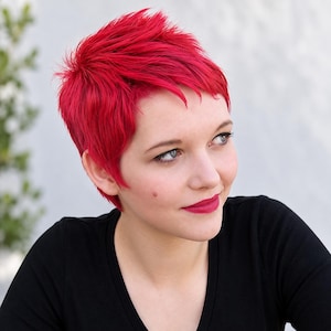 May include: A person with short, vibrant red hair styled in a textured pixie cut. The individual has a fair complexion, dark eyebrows, and is wearing a plain black v-neck shirt. The hair is bright, saturated red with visible layers and spikes.