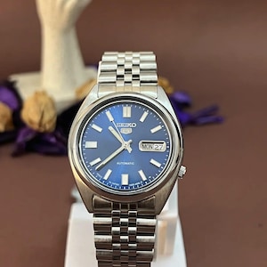 May include: A silver-tone Seiko 5 automatic wristwatch featuring a round blue dial with luminous hour markers, silver hands, and a day-date window at the three o'clock position. The watch includes a stainless steel link bracelet with a fold-over clasp.