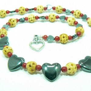 May include: A necklace featuring carved beige beads, red beads, and dark gray heart-shaped beads. The necklace has a silver heart-shaped charm and a silver clasp. The beads are strung together to form a unique jewelry piece.