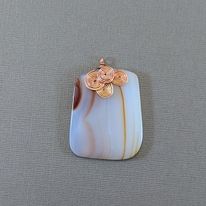 May include: A rectangular pendant with a polished, light-colored stone featuring brown and white veining. The pendant is adorned with copper wire swirls at the top. A small copper loop is at the top for hanging.