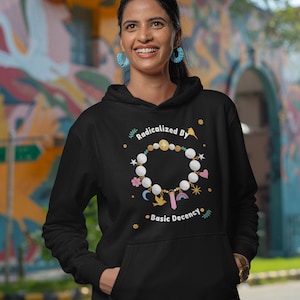 Pode incluir: Uma camisola preta com capuz apresentando uma estampa gráfica de uma pulseira circular de contas com berloques, incluindo um pássaro, um cristal e um arco-íris. O texto "Radicalized By" aparece acima da pulseira e "Basic Decency" abaixo.