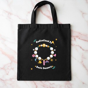 Puede incluir: Un bolso tote negro con un gráfico de una pulsera circular de cuentas con varios dijes, incluyendo un pájaro, un arcoíris, un cristal, una luna y un corazón. El texto "Radicalized By Basic Decency" está impreso en blanco sobre el diseño.